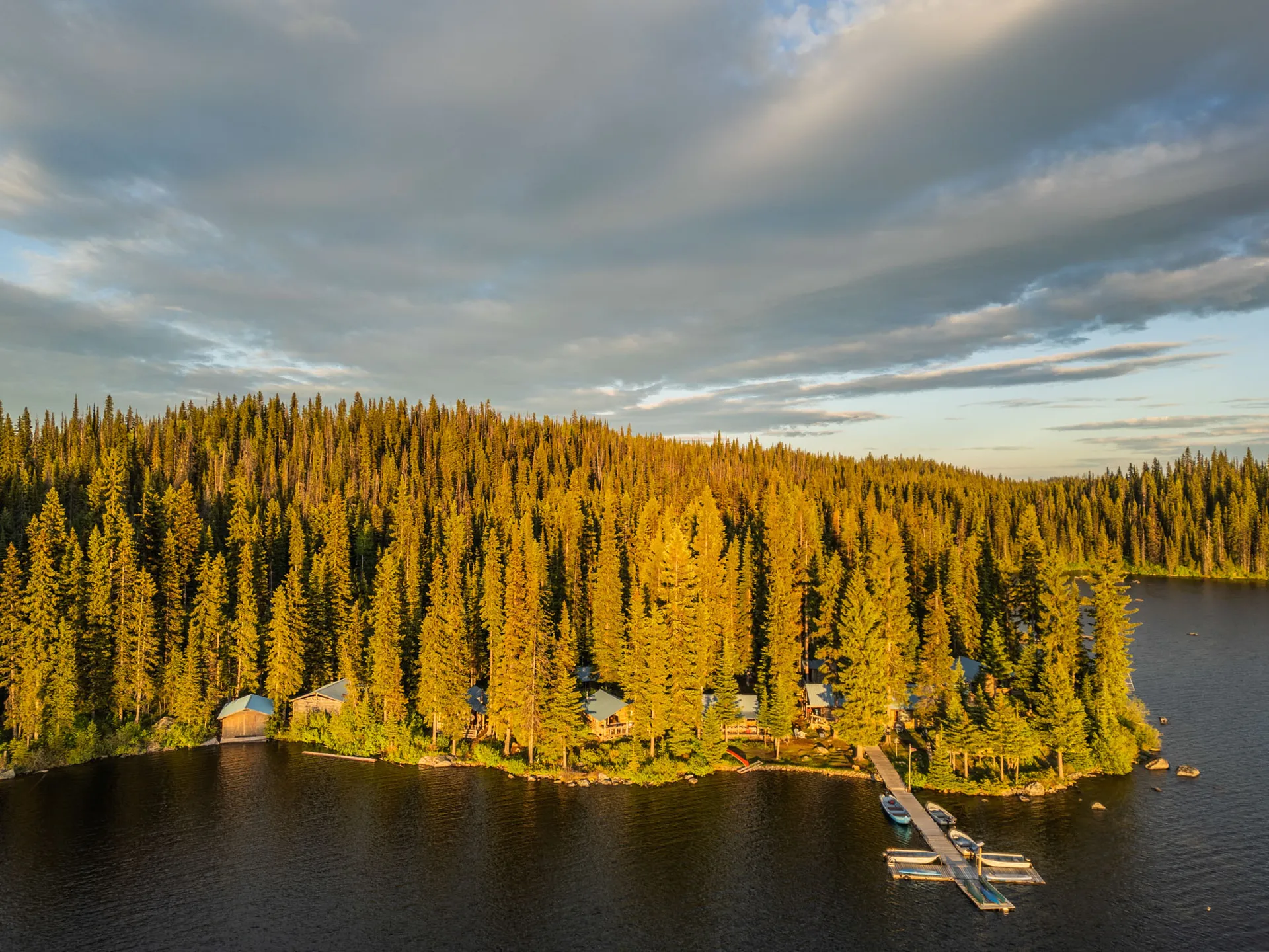 Aerial view of Caverhill's log cabins along a forested lakeshore, with a dock and boats in golden evening light