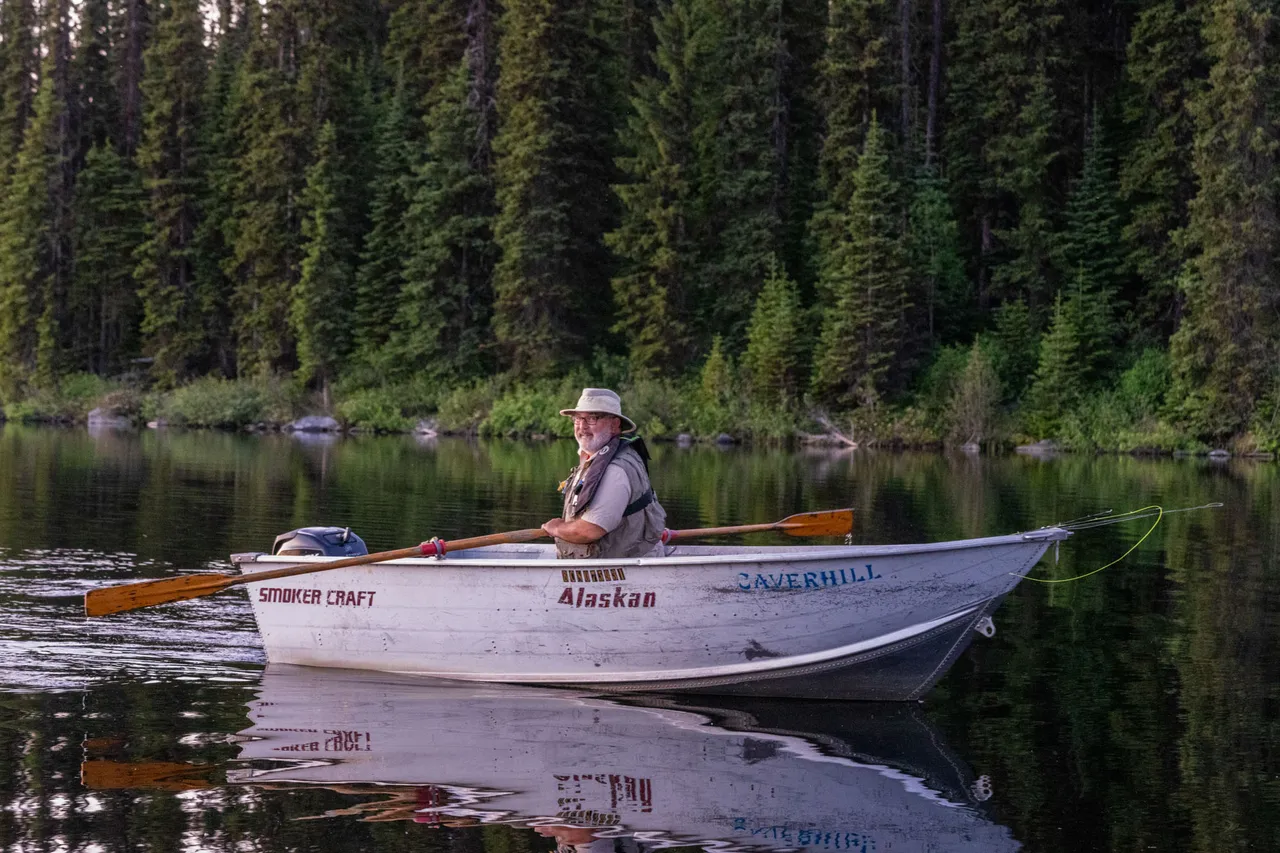 An angler in a white Caverhill dory on still water, the forested shoreline reflected behind