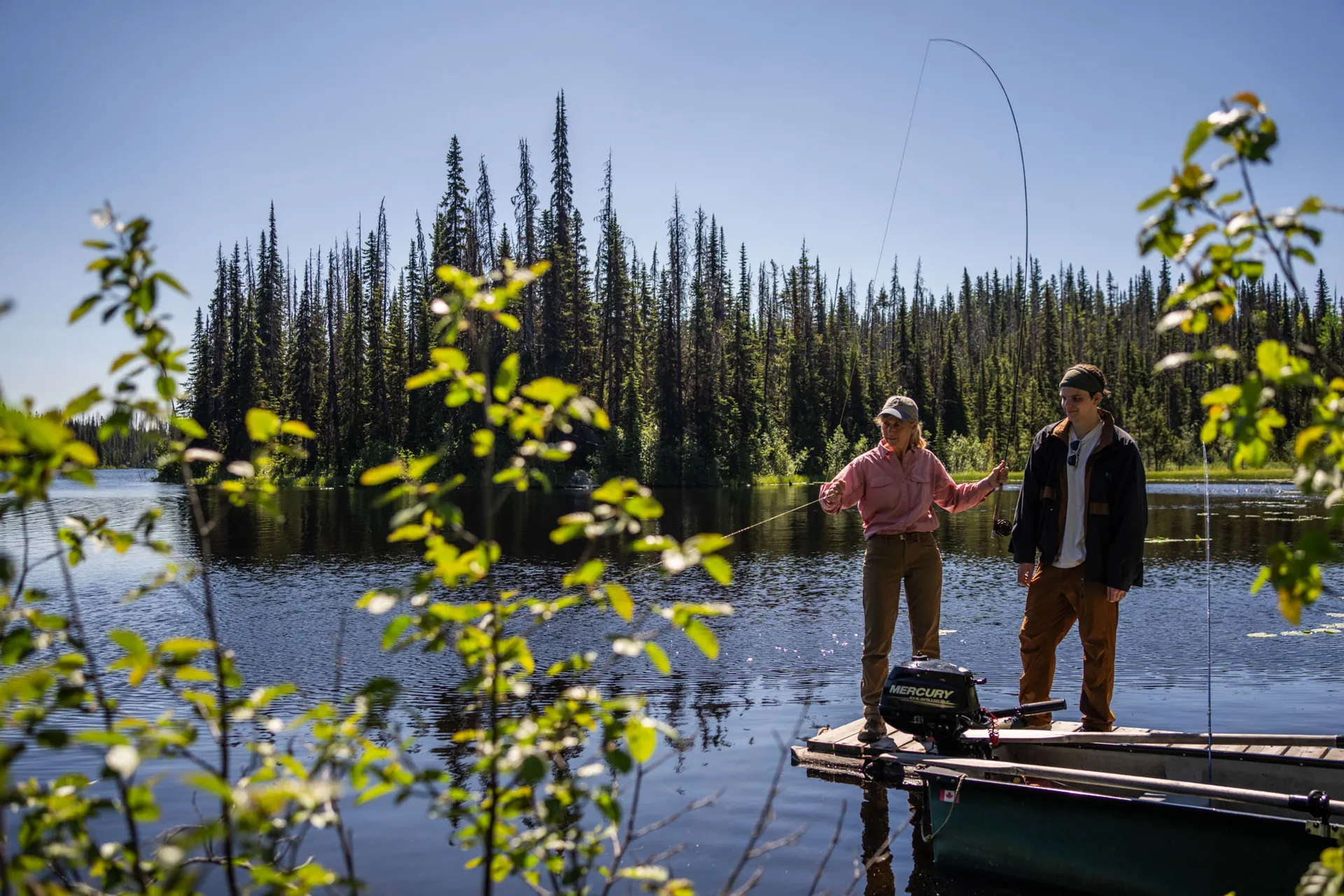 Two anglers fly casting from a boat dock, a spruce-lined shore across the lake