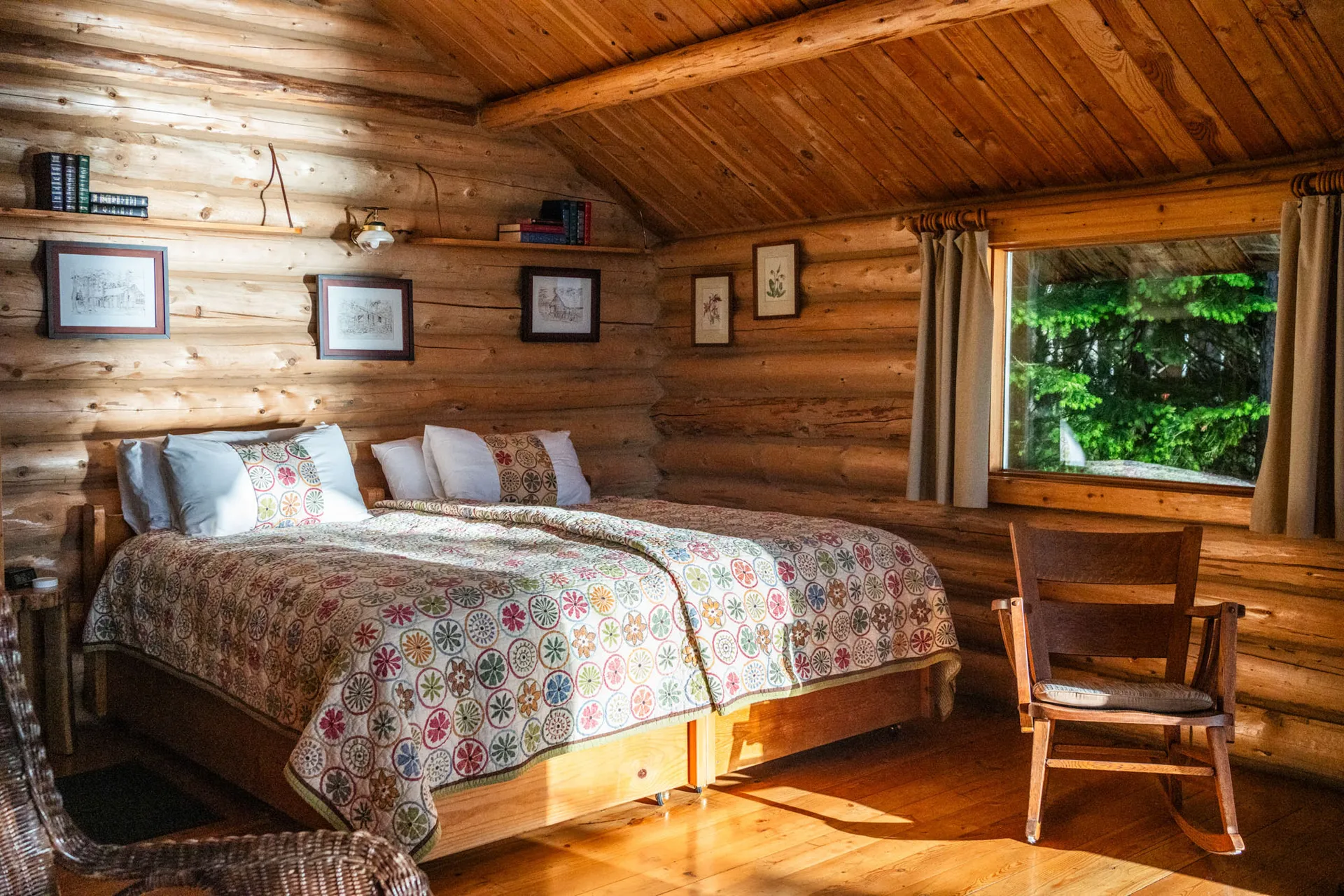 Log cabin bedroom with a quilted bed, framed pictures on the timber wall and a window onto the forest