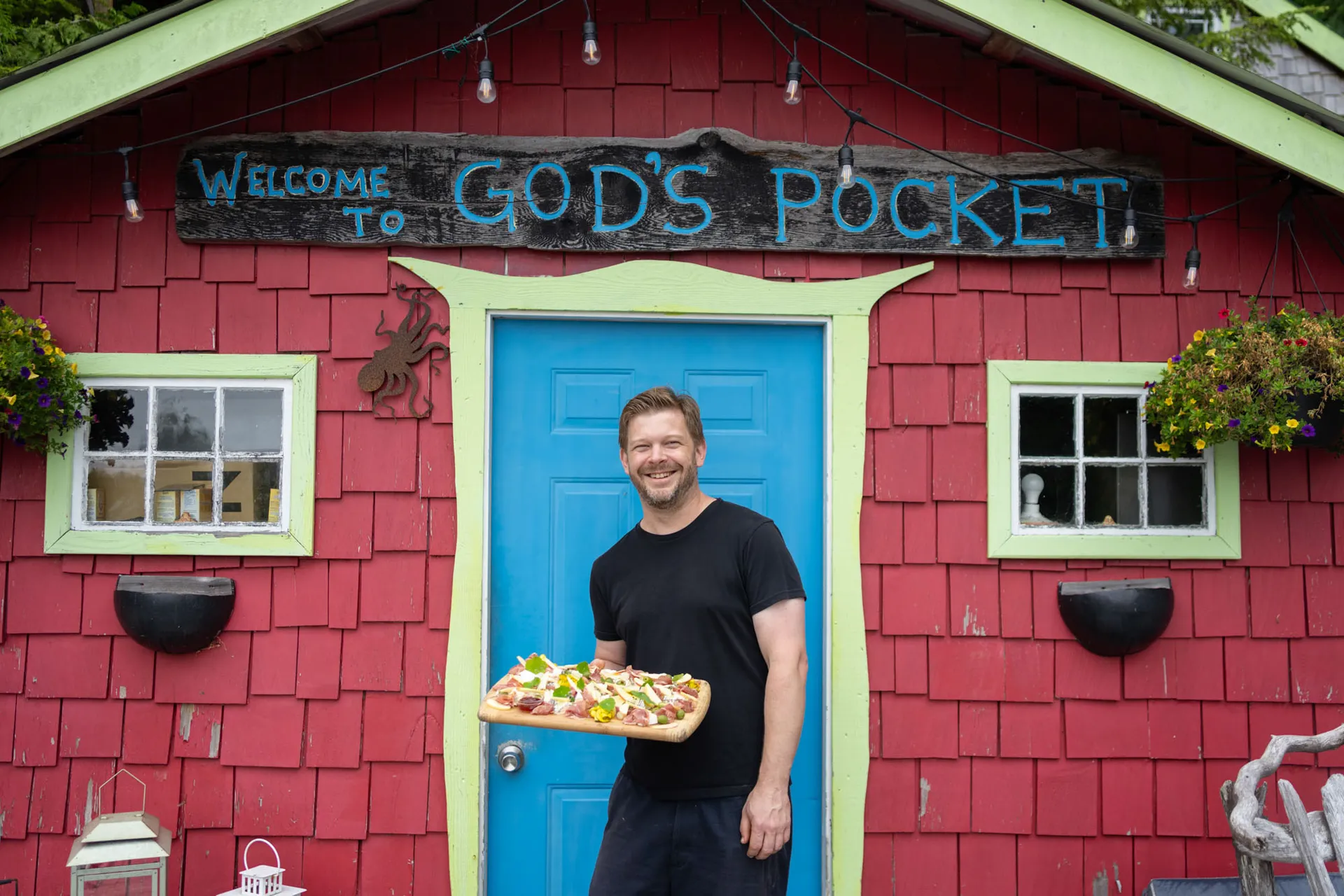 Host standing at the red cabin entrance beneath a hand-painted Welcome to God's Pocket sign, holding a board of pizza