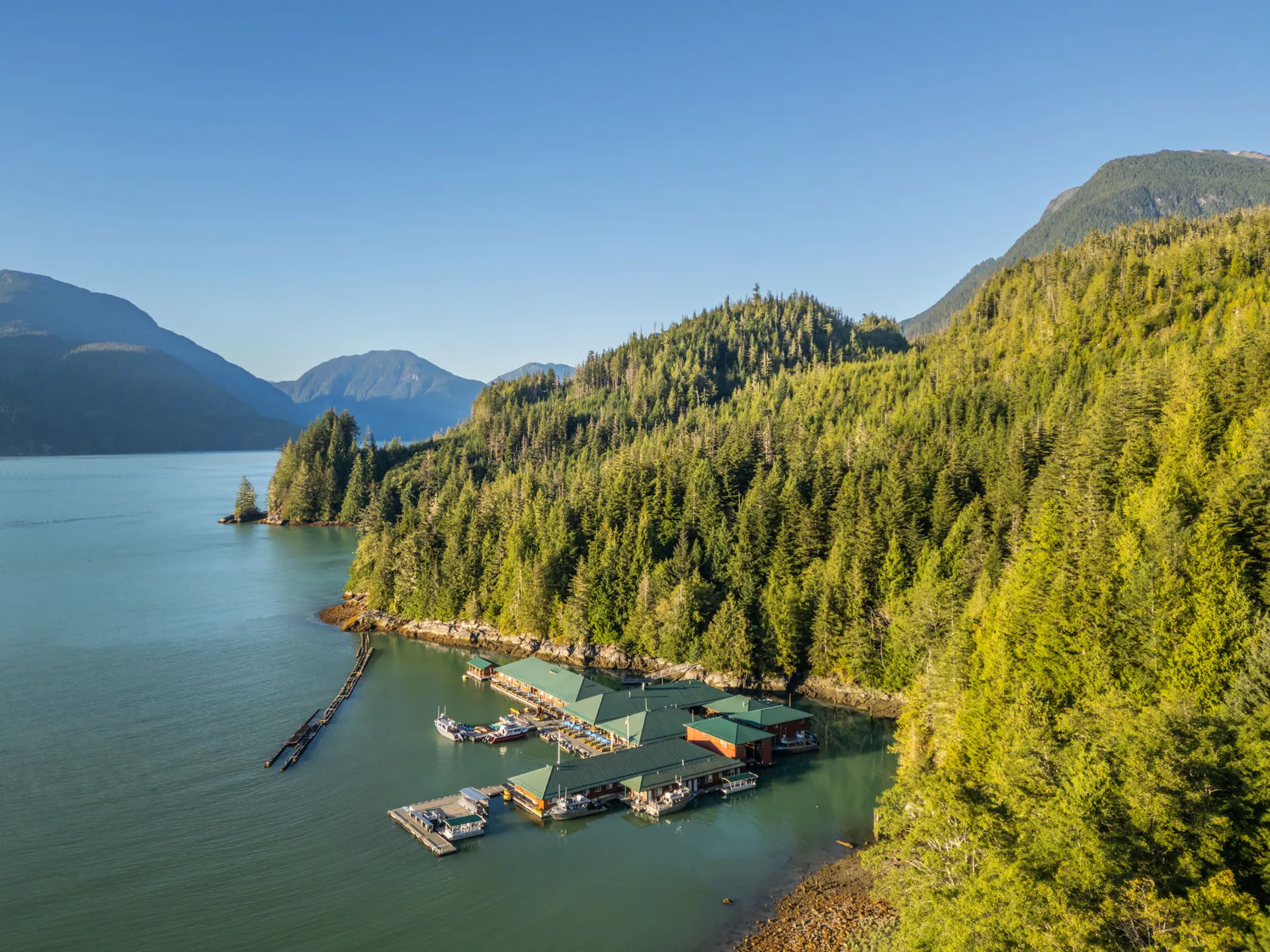 Aerial view along Knight Inlet showing the green-roofed floating lodge tied against the forested shoreline, mountains beyond