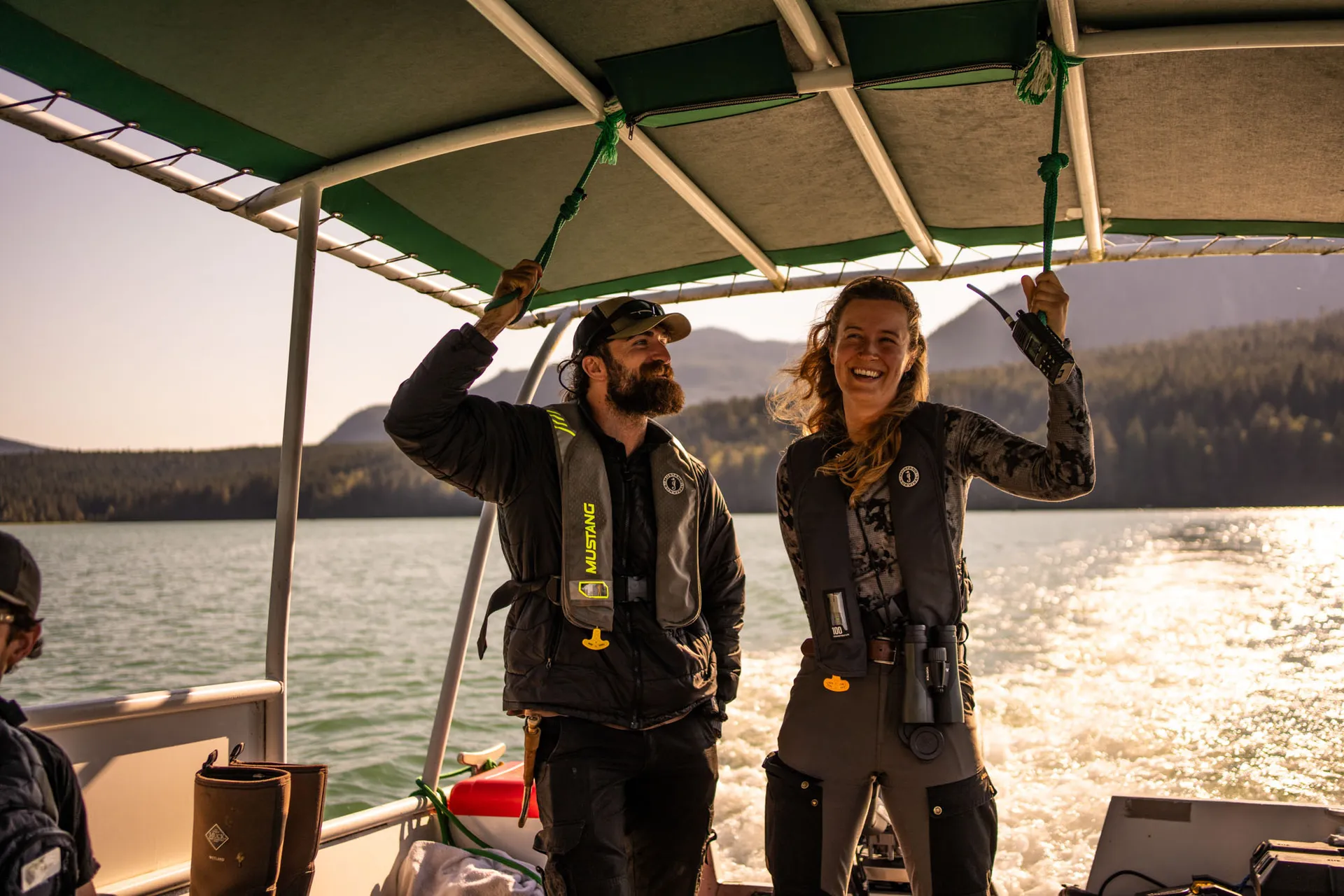 Two guides in floater jackets laughing aboard a covered viewing boat under low evening sun
