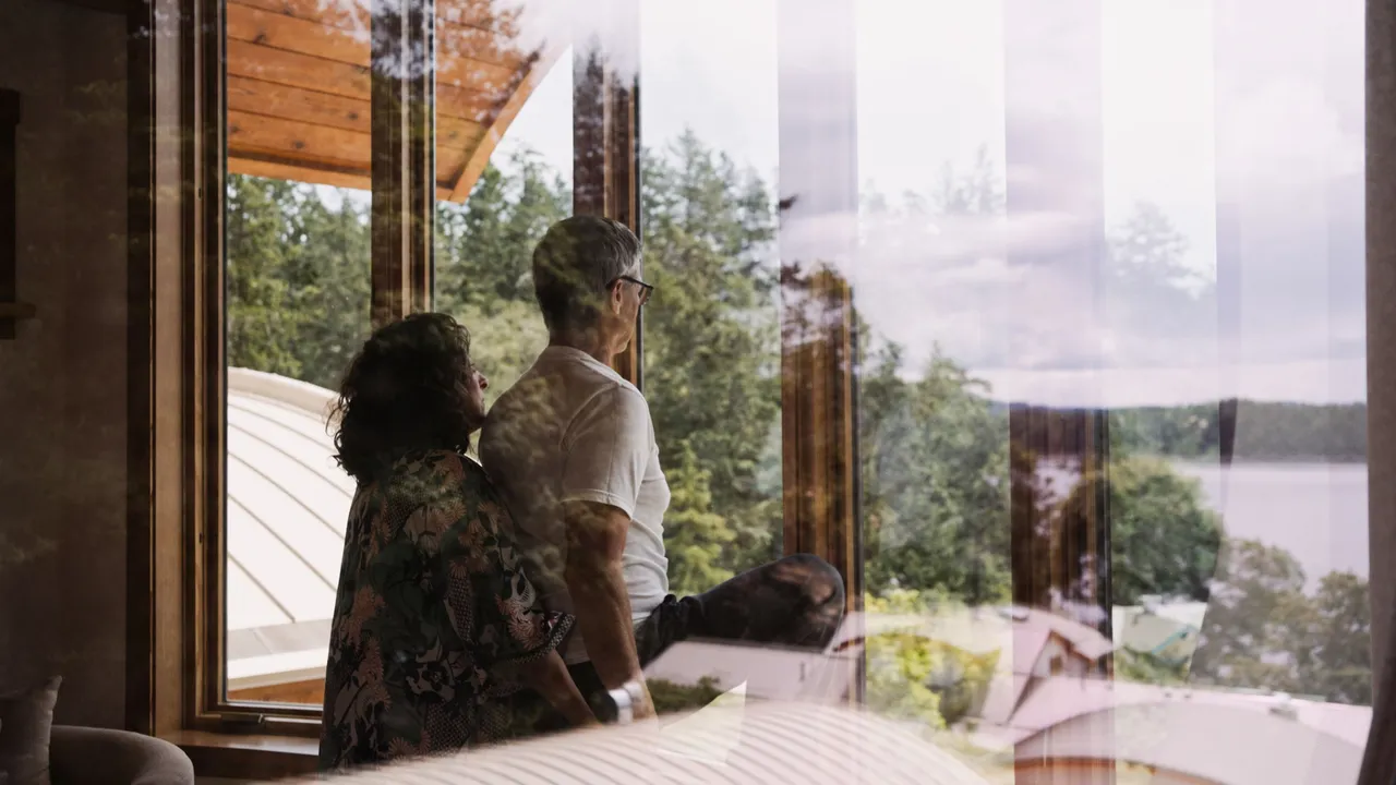 Two guests looking out through tall timber-framed windows toward the water and distant islands