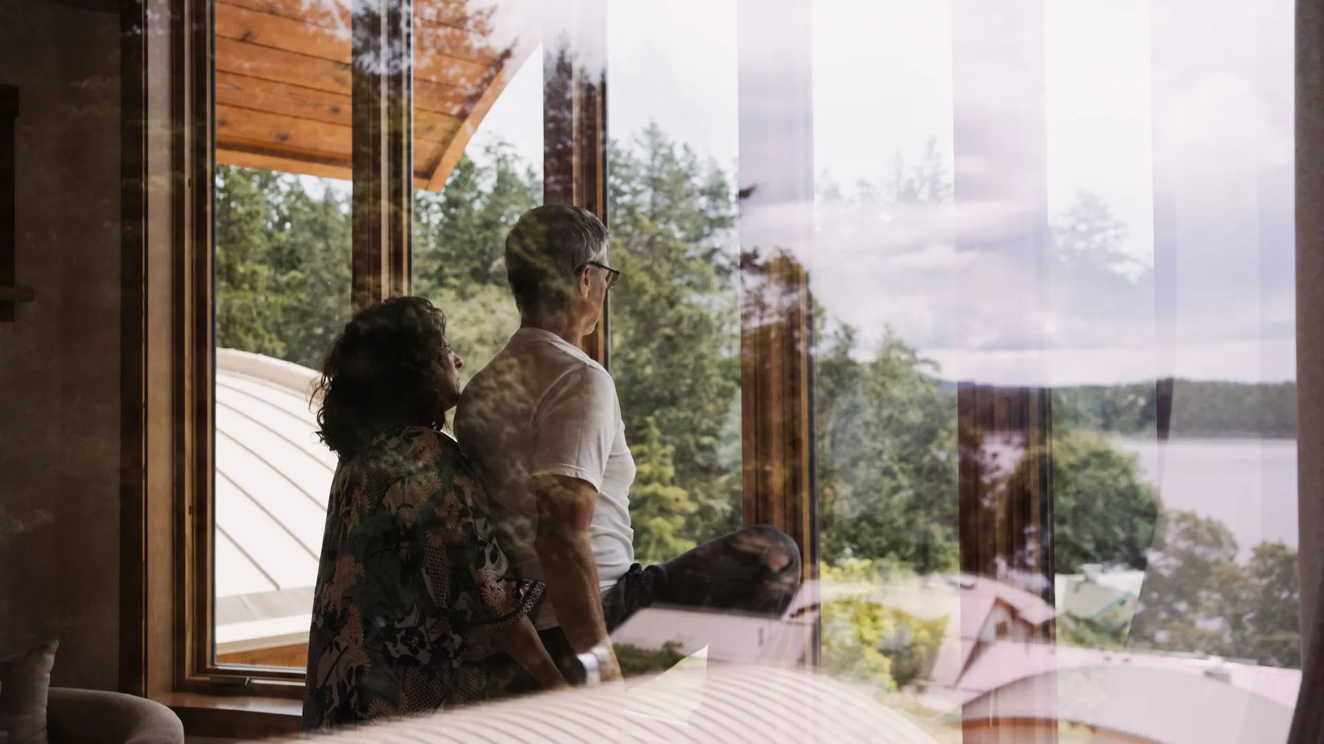Two guests looking out through tall timber-framed windows toward the water and distant islands