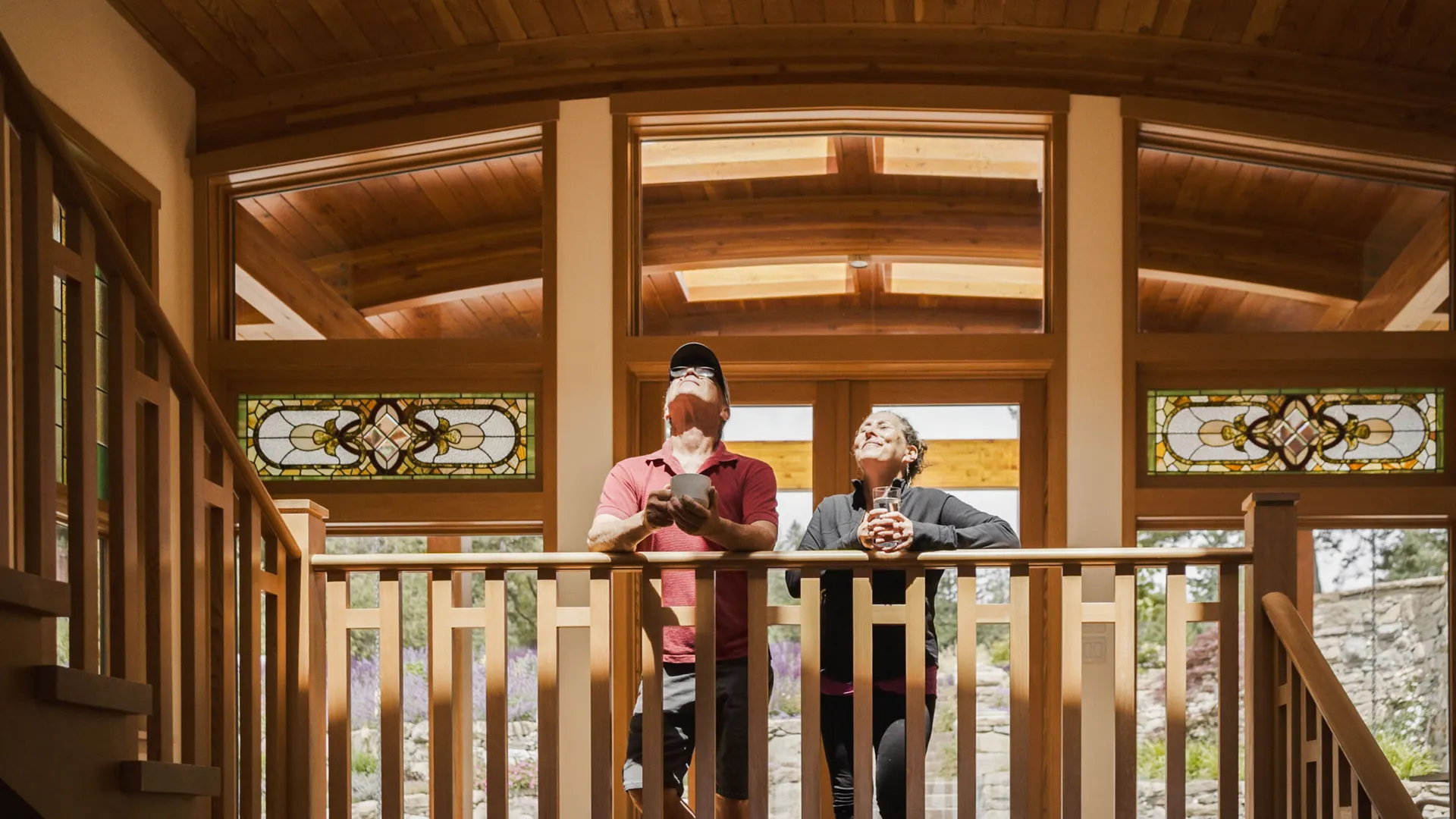 Two guests leaning on an interior balcony rail beneath a curved cedar ceiling and stained-glass panels