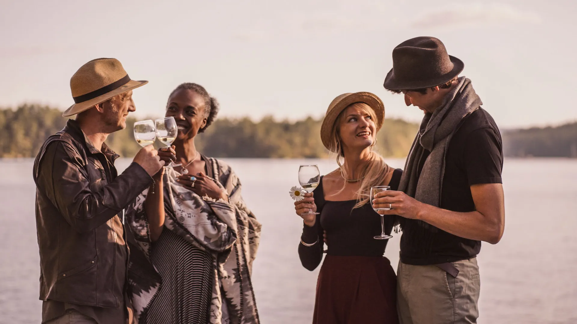 Four guests standing with glasses of wine by the water at golden hour