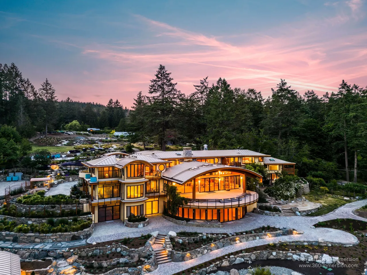 Aerial view of Lightwater Cove's curved timber-and-glass guesthouse lit from within at dusk, ringed by terraced stone gardens