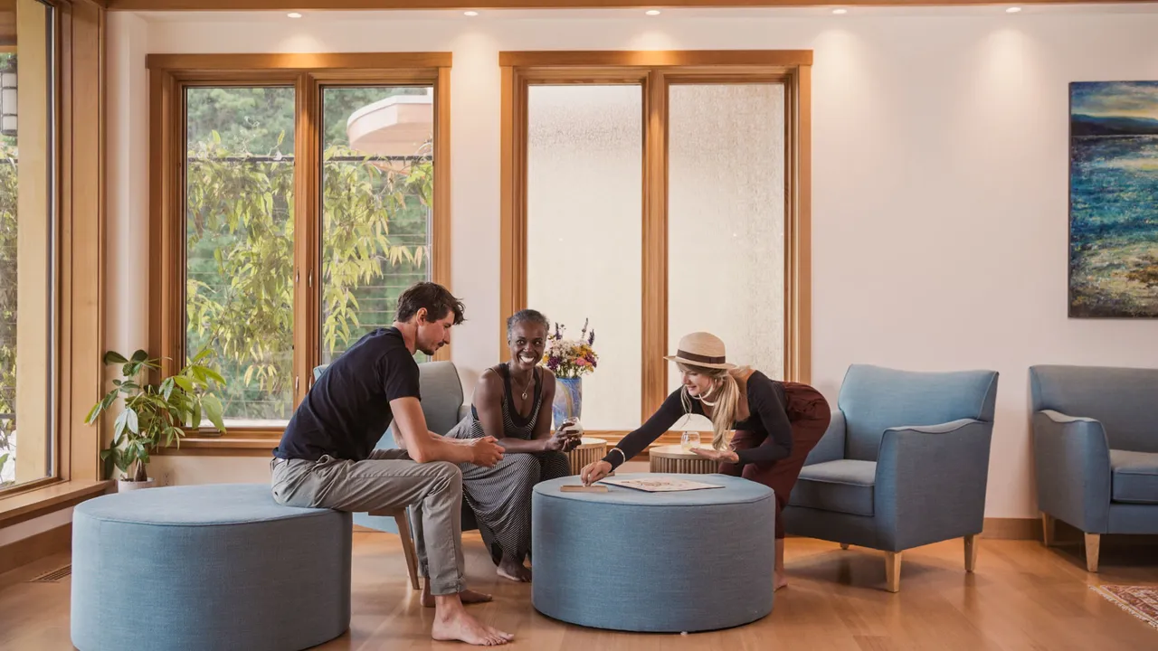 Guests seated on round upholstered ottomans in a light-filled lounge with timber-framed windows
