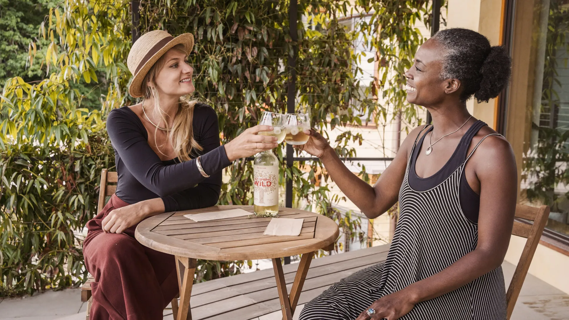 Two guests toasting with drinks at a small table on a vine-shaded terrace