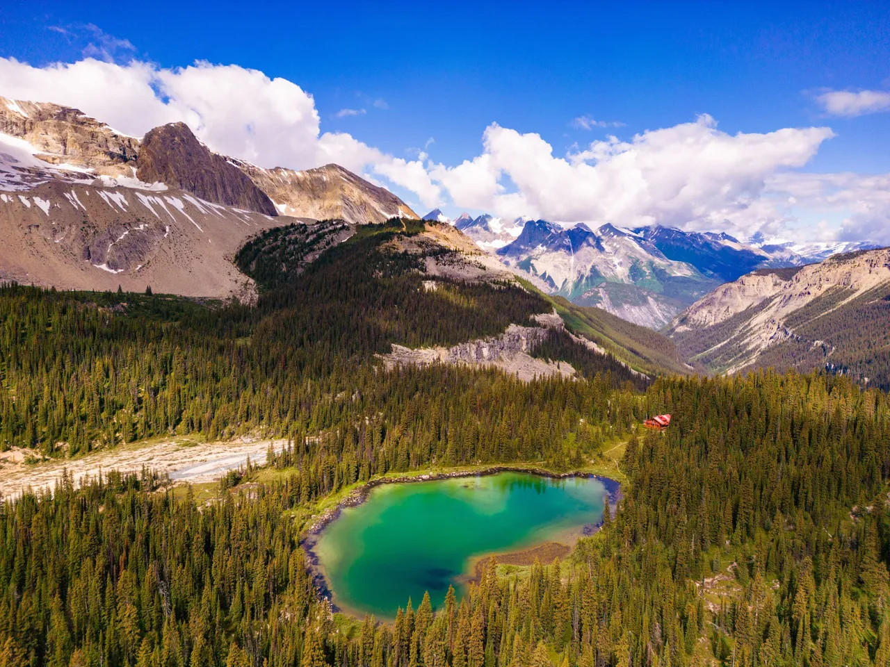 Aerial view of the lodge above a turquoise alpine tarn, with glaciated Rocky Mountain peaks beyond