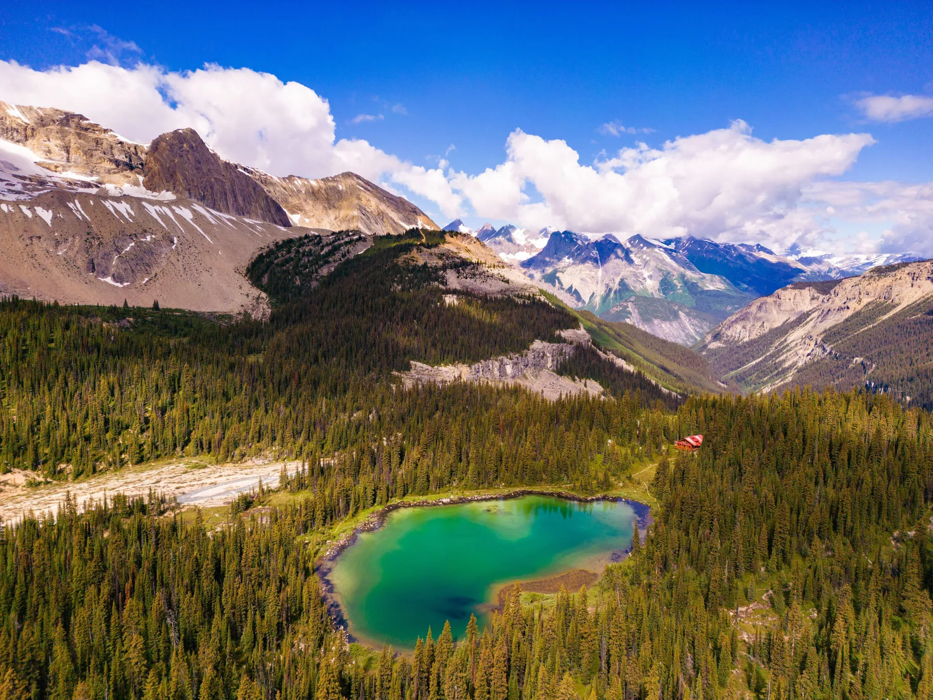 Aerial view of the lodge above a turquoise alpine tarn, with glaciated Rocky Mountain peaks beyond
