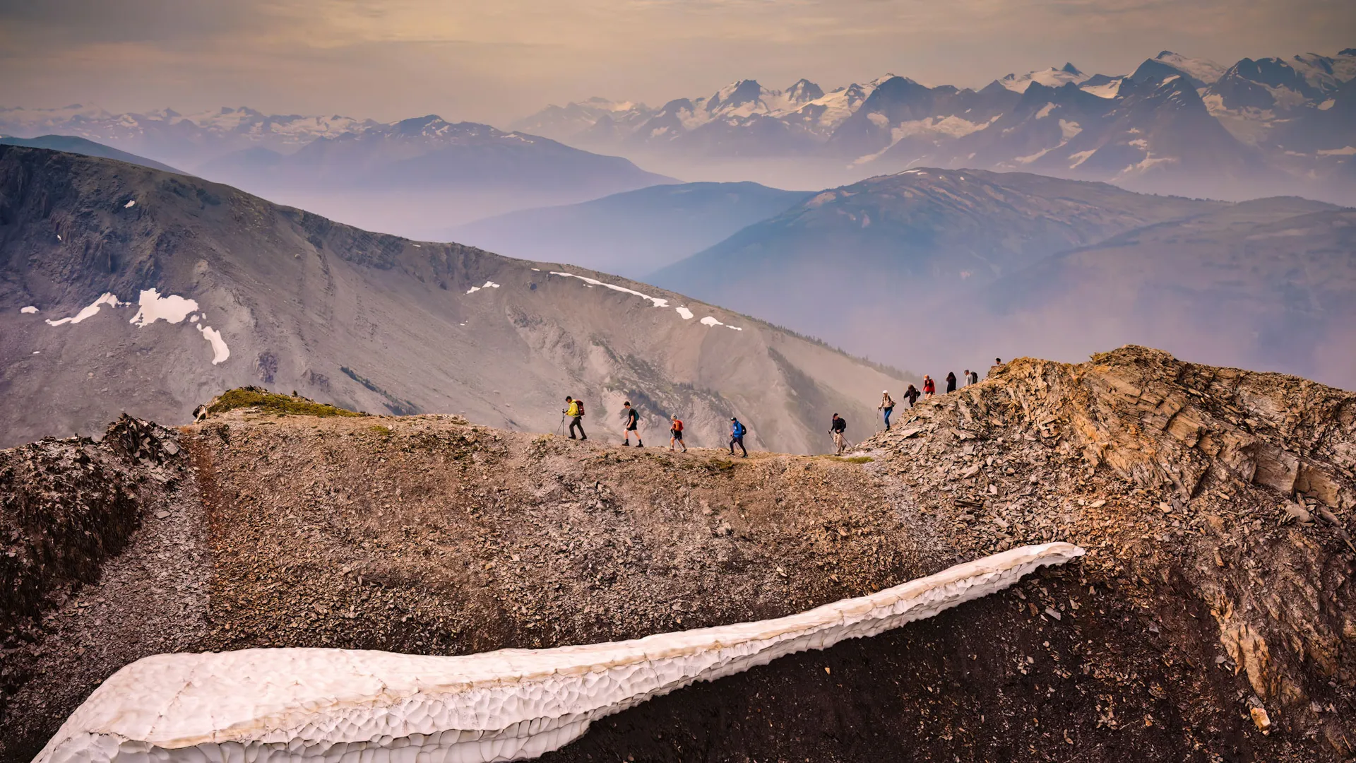 A line of hikers strung along a high ridge above a snow cornice, glaciated peaks behind