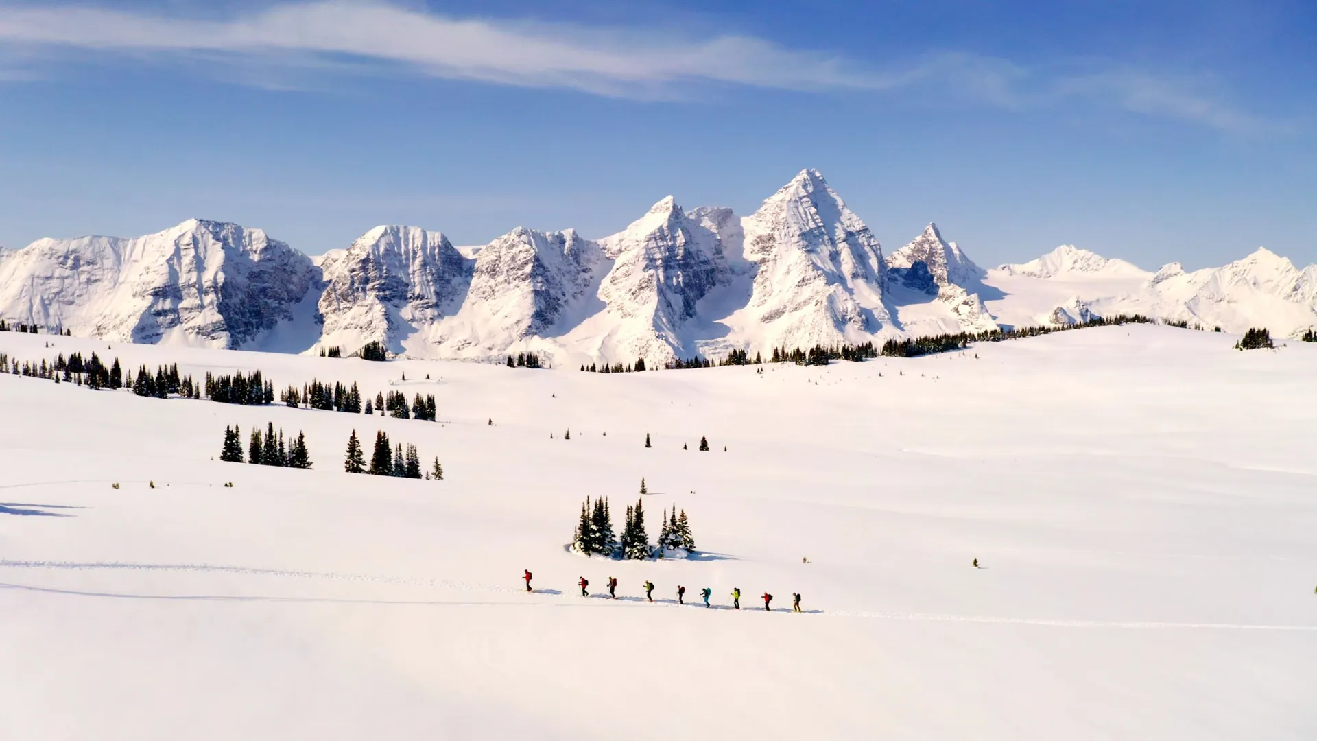 A line of ski tourers skinning across an open snowfield below a wall of snow-covered peaks