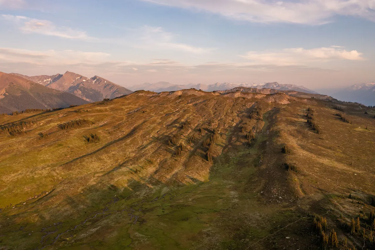 Late sun raking across a broad alpine ridge of meadow and scree, mountains fading into haze