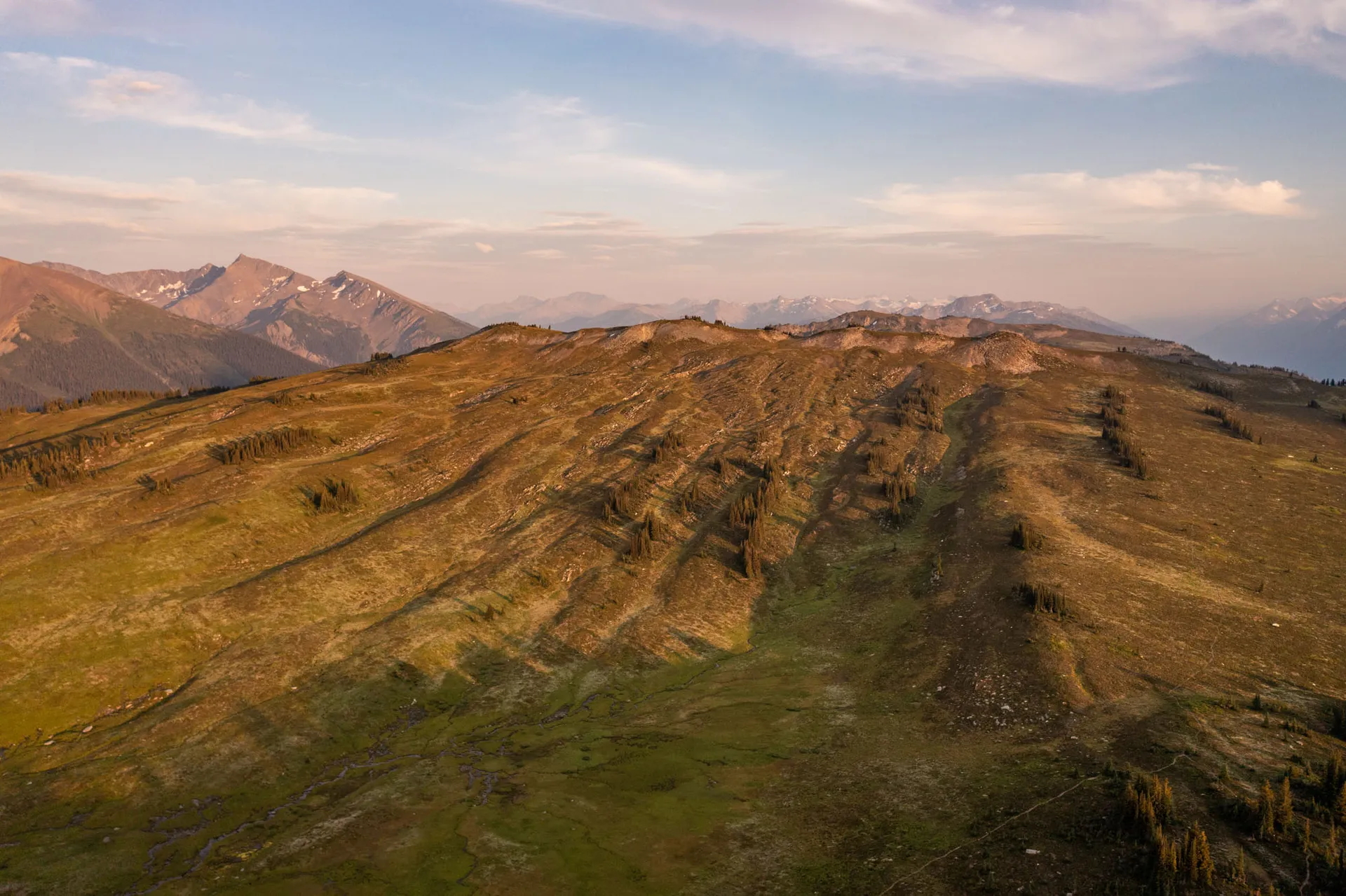 Late sun raking across a broad alpine ridge of meadow and scree, mountains fading into haze