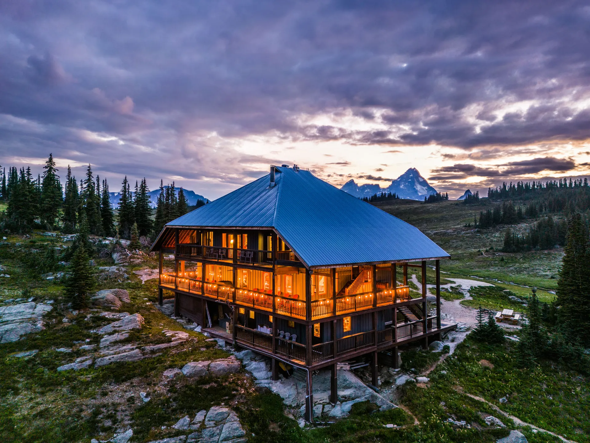 Purcell Mountain Lodge, a three-storey timber lodge with a blue roof, lit at sunset with jagged peaks behind