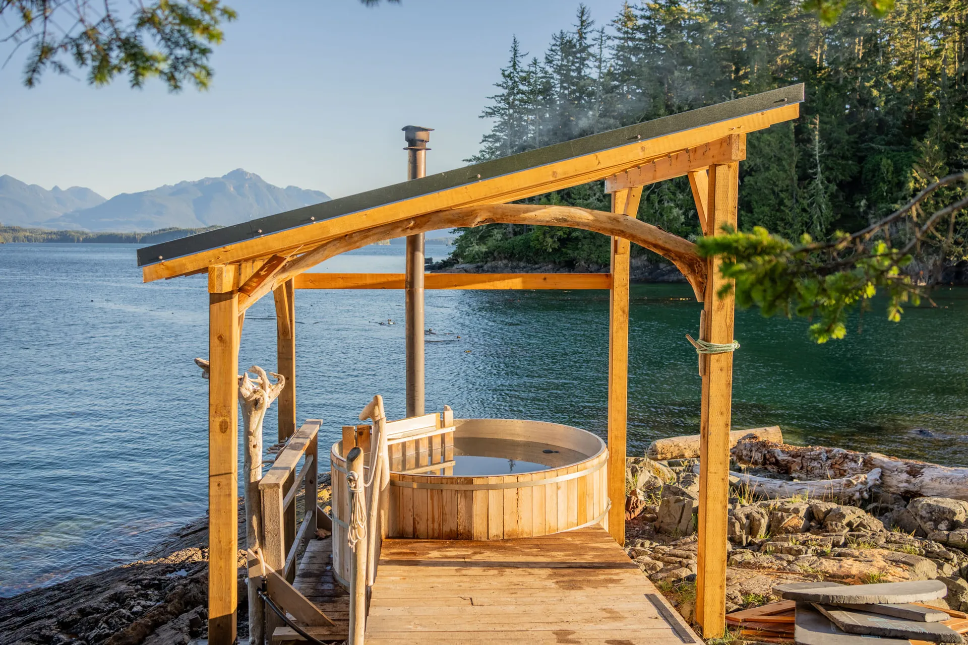 Cedar hot tub under a timber shelter at the water's edge, looking across the channel to distant mountains