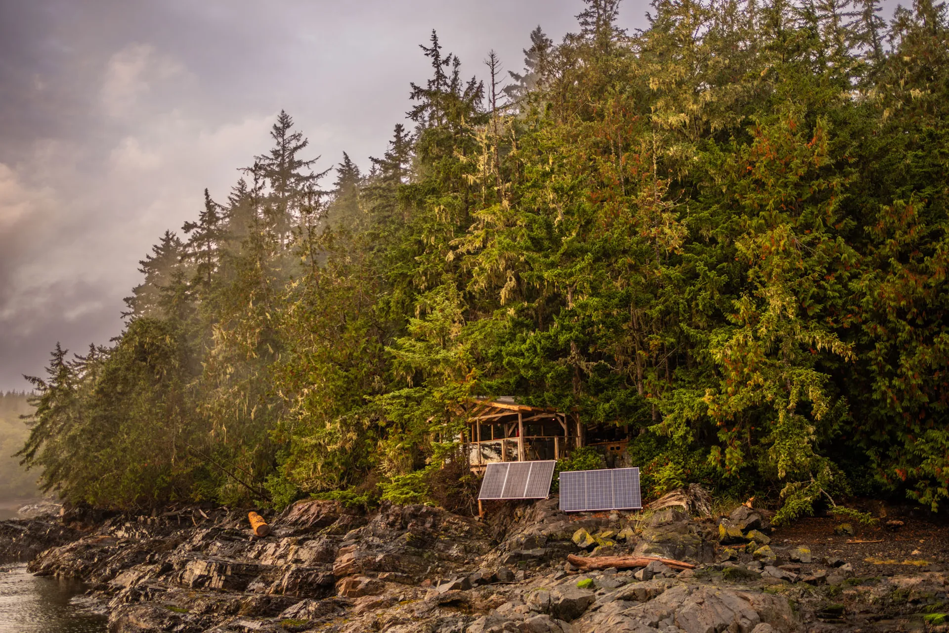 The base camp's timber cook shelter and solar panels set among trees above a rocky shore