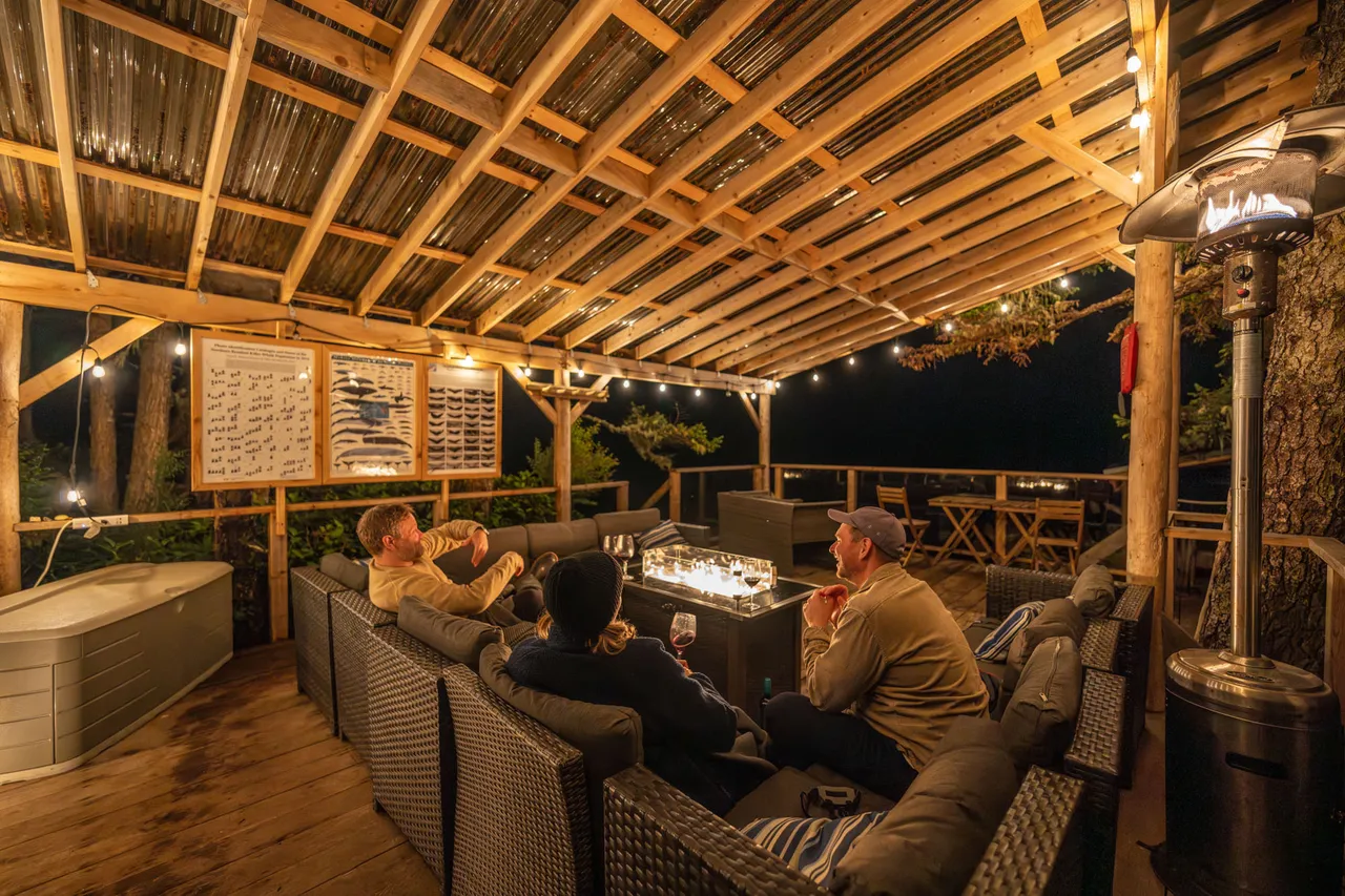 Guests around a lit fire table under a covered deck in the evening, with a hot tub alongside