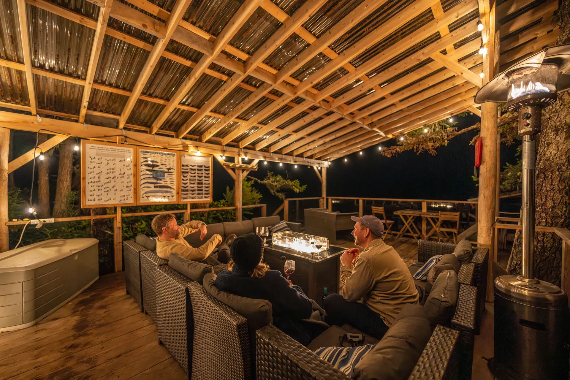 Guests around a lit fire table under a covered deck in the evening, with a hot tub alongside