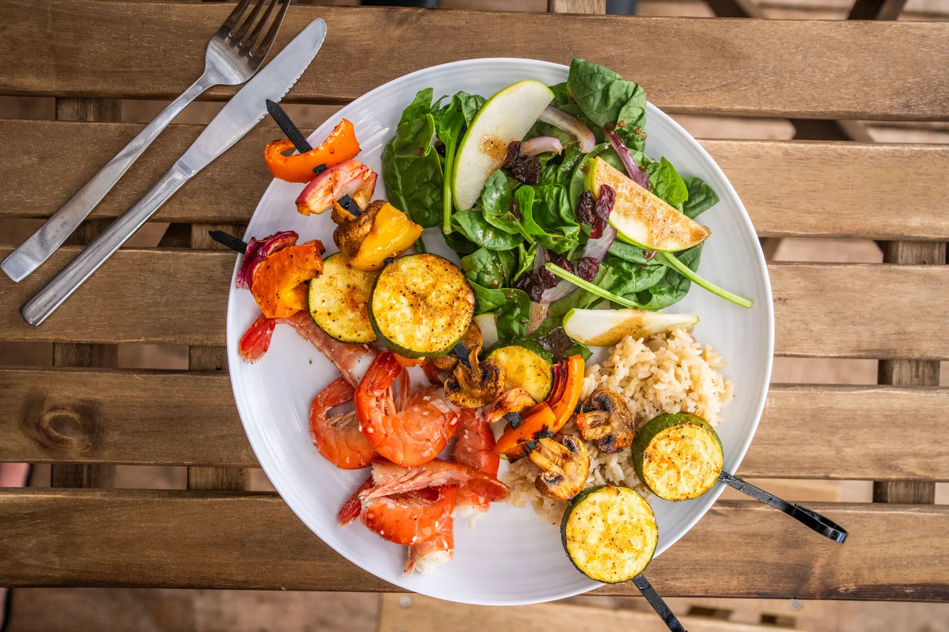 Plate of grilled prawns, vegetable skewers, rice and spinach salad on a wooden table
