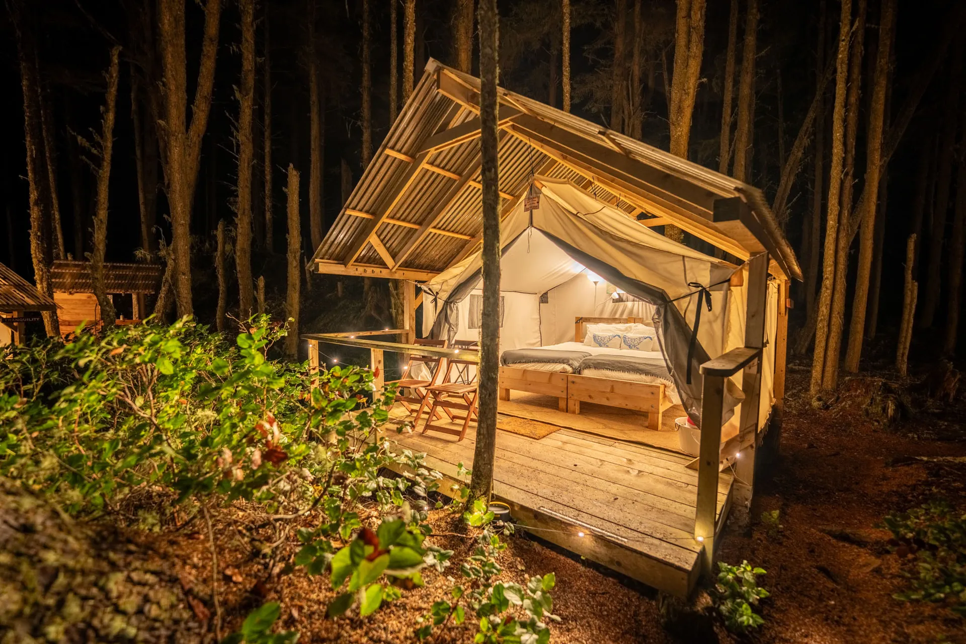 Safari tent lit from within at night, its bed and deck visible under a peaked timber frame