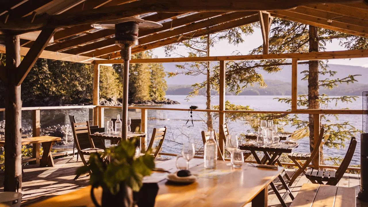 Long dining table set with glassware under an open timber shelter overlooking the water