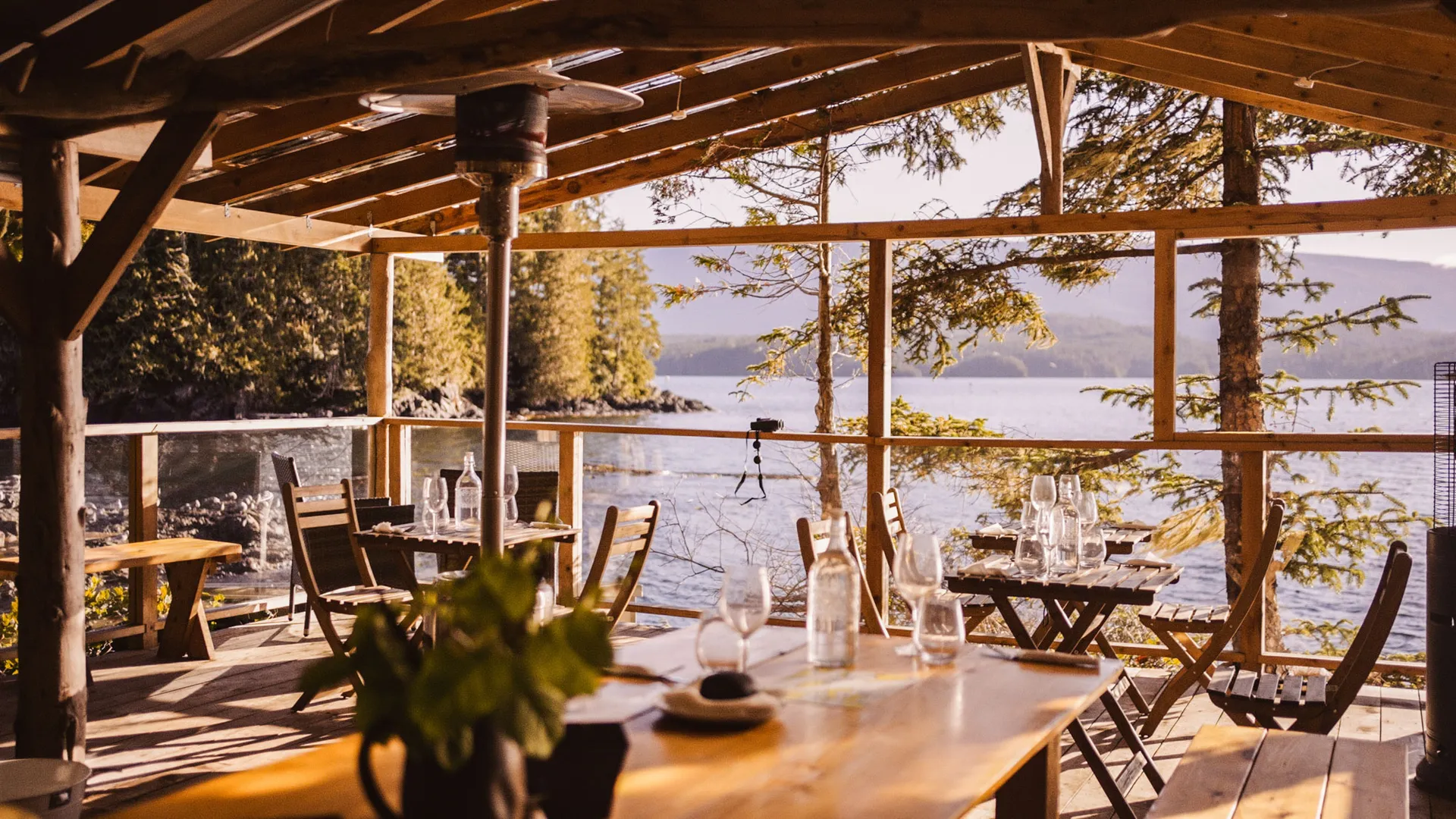 Long dining table set with glassware under an open timber shelter overlooking the water
